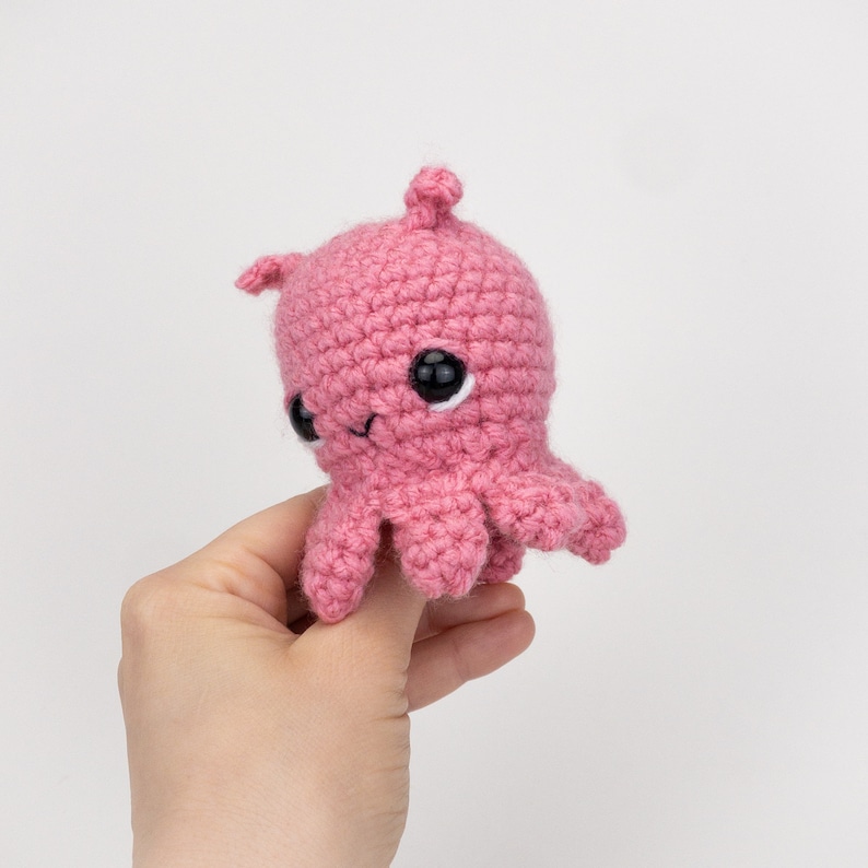 Puede incluir: Un pulpo de crochet rosa con ojos negros. El pulpo tiene una peque&ntilde;a boca sonriente.