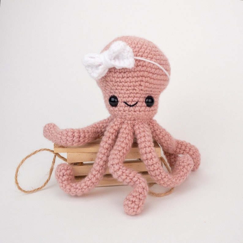 May include: A pink crocheted octopus with a white bow on its head. The octopus is sitting on a wooden crate.