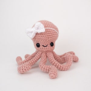 May include: A pink crocheted octopus with a white bow on its head. The octopus has eight tentacles and a smiling face.
