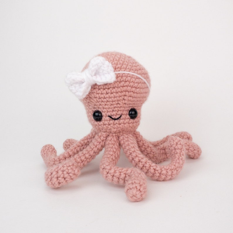 May include: A pink crocheted octopus with a white bow on its head. The octopus has eight tentacles and a smiling face.