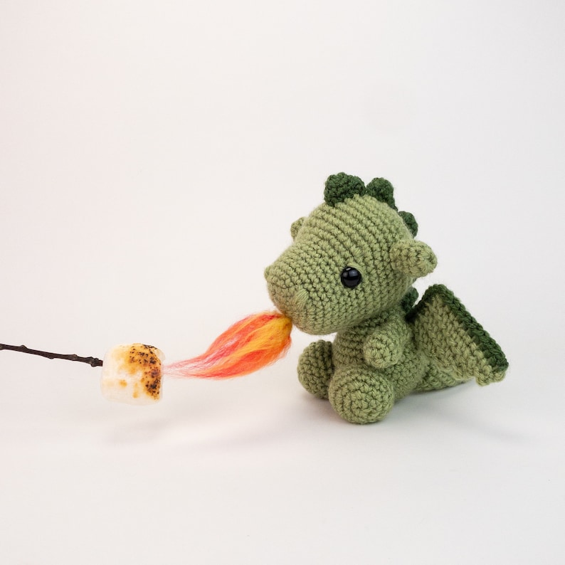 May include: A green crocheted dragon with a red and yellow yarn flame coming out of its mouth. The dragon is sitting on a white surface.