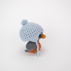 May include: A handmade crochet bird figurine wearing a light blue hat with a pom-pom and ear flaps. The bird has a gray back, orange chest, and yellow beak. The figurine is a charming, whimsical creation.