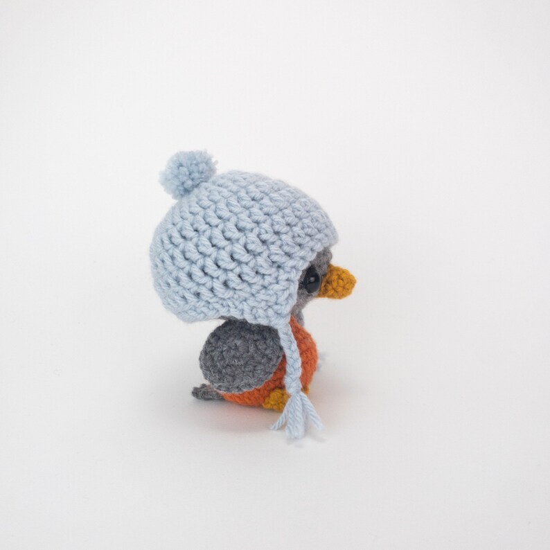 May include: A handmade crochet bird figurine wearing a light blue hat with a pom-pom and ear flaps. The bird has a gray back, orange chest, and yellow beak. The figurine is a charming, whimsical creation.