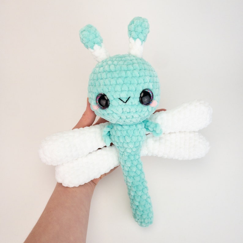 May include: A crocheted dragonfly toy with a light blue body, white wings, and black eyes. The dragonfly has a cute, cartoon-like face.