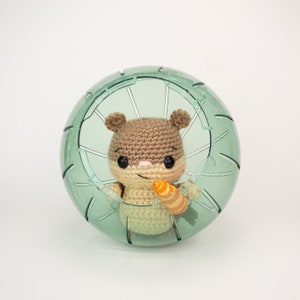 May include: A brown and beige crocheted hamster holding a small orange carrot, sitting inside a clear plastic hamster ball.