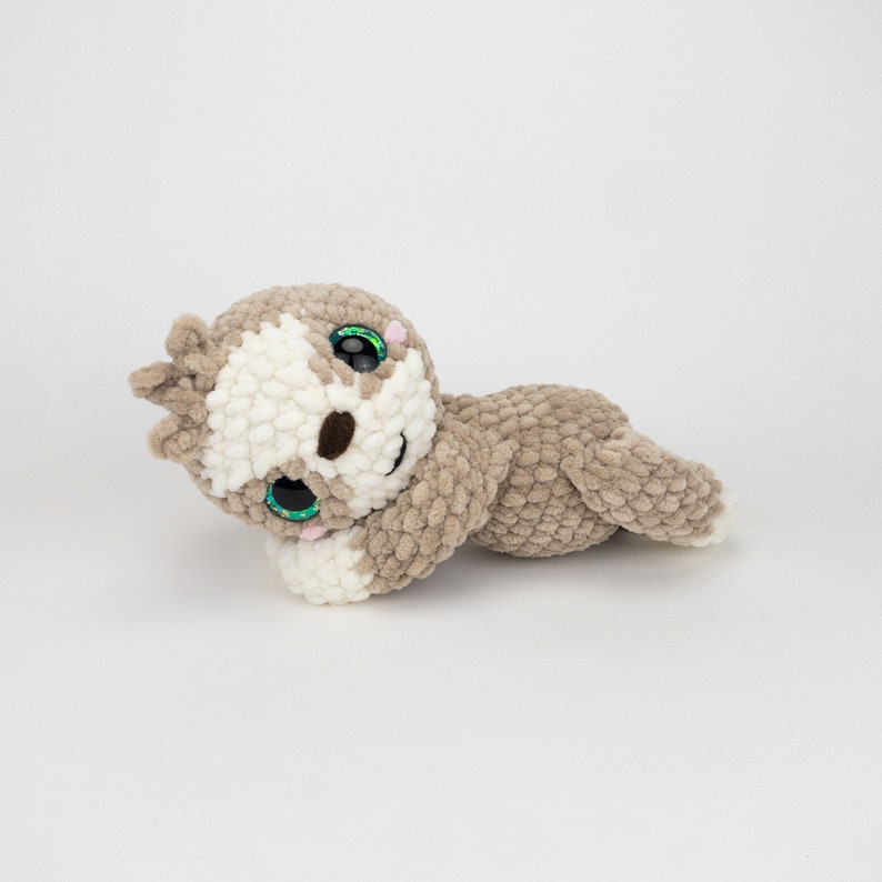 May include: A crocheted sloth plush toy with brown and white fur, green eyes, and a pink nose. The sloth is lying on its side with its head resting on its arm.
