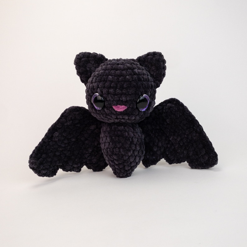 K&ouml;nnte beinhalten: Ein schwarzes, geh&auml;keltes Fledermaus-Pl&uuml;schtier mit gro&szlig;en lila Augen und einer rosa Nase. Die Fledermaus hat gro&szlig;e Fl&uuml;gel und einen kleinen K&ouml;rper.