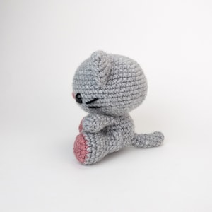 Puede incluir: Un juguete de crochet de gato gris con almohadillas de patas rosas. El gato est&aacute; sentado con la cola enroscada alrededor de su cuerpo.