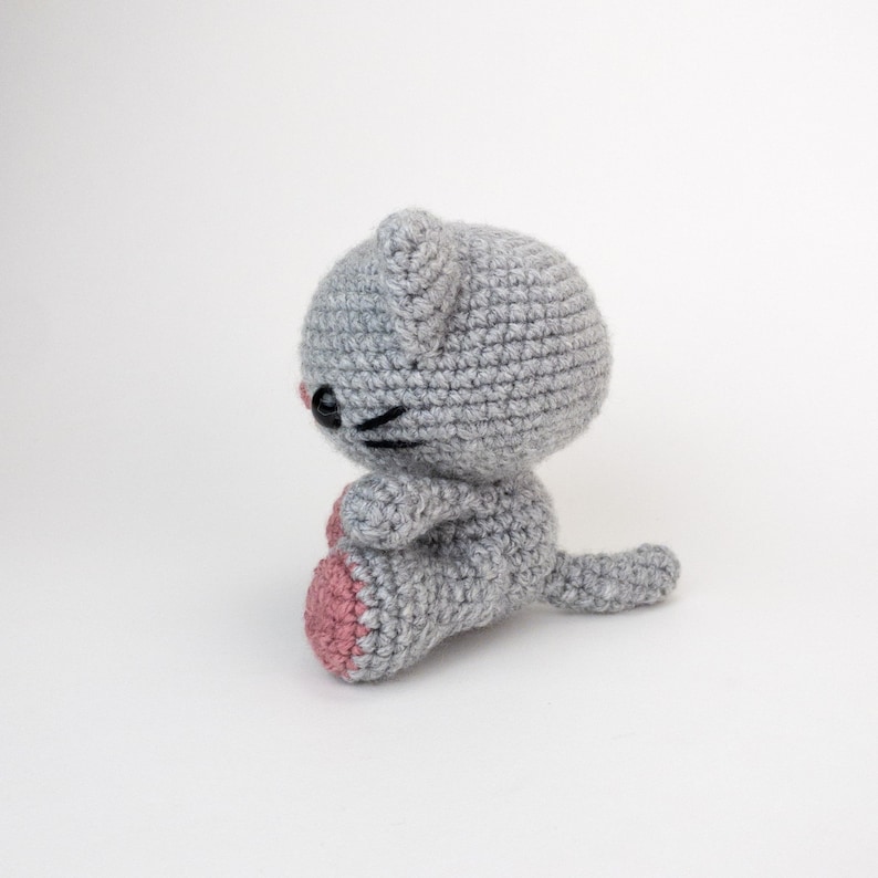 Puede incluir: Un juguete de crochet de gato gris con almohadillas de patas rosas. El gato est&aacute; sentado con la cola enroscada alrededor de su cuerpo.