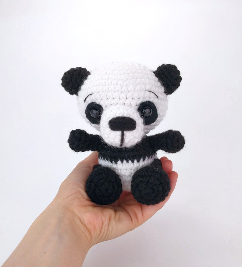 May include: A small crocheted panda bear toy with black and white fur, black eyes, and a black nose. The panda is sitting on a hand.