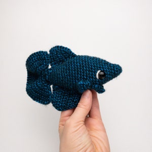 May include: A crocheted blue betta fish with white eyes. The fish is being held in a hand.