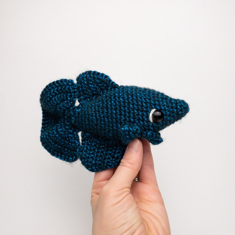 May include: A crocheted blue betta fish with white eyes. The fish is being held in a hand.