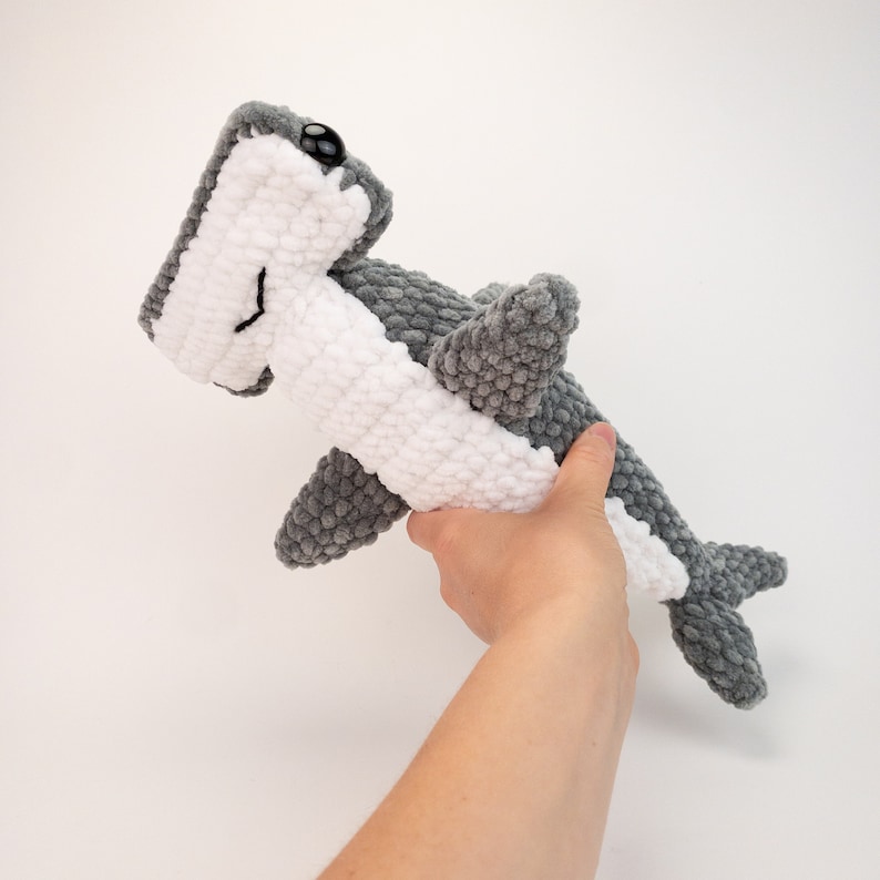 May include: A crocheted hammerhead shark plush toy with white and gray yarn. The plush toy has black eyes and a smiling mouth.