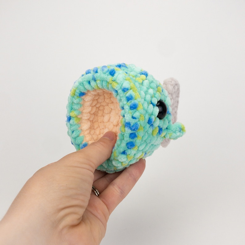 May include: A crocheted plush toy shaped like a narwhal with a blue and green speckled body, a pink horn, and a black eye.