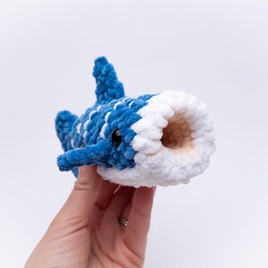 May include: A blue and white crocheted whale plush toy with a large open mouth. The whale has black eyes and a white belly.