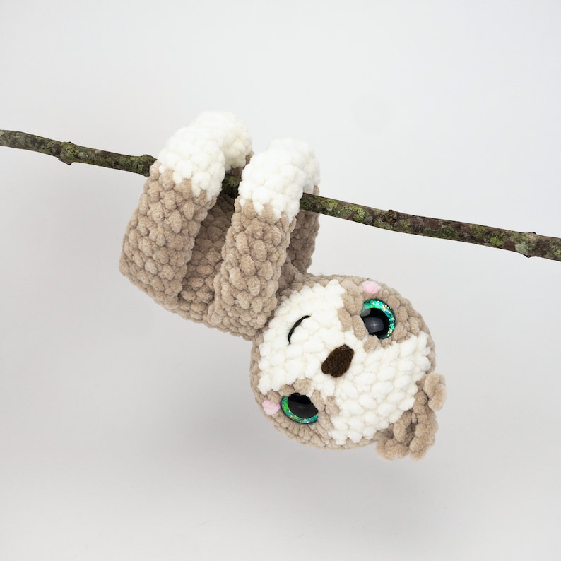 May include: A crocheted sloth toy with brown and white fur hanging upside down from a branch. The sloth has large green eyes with glitter and a pink nose.