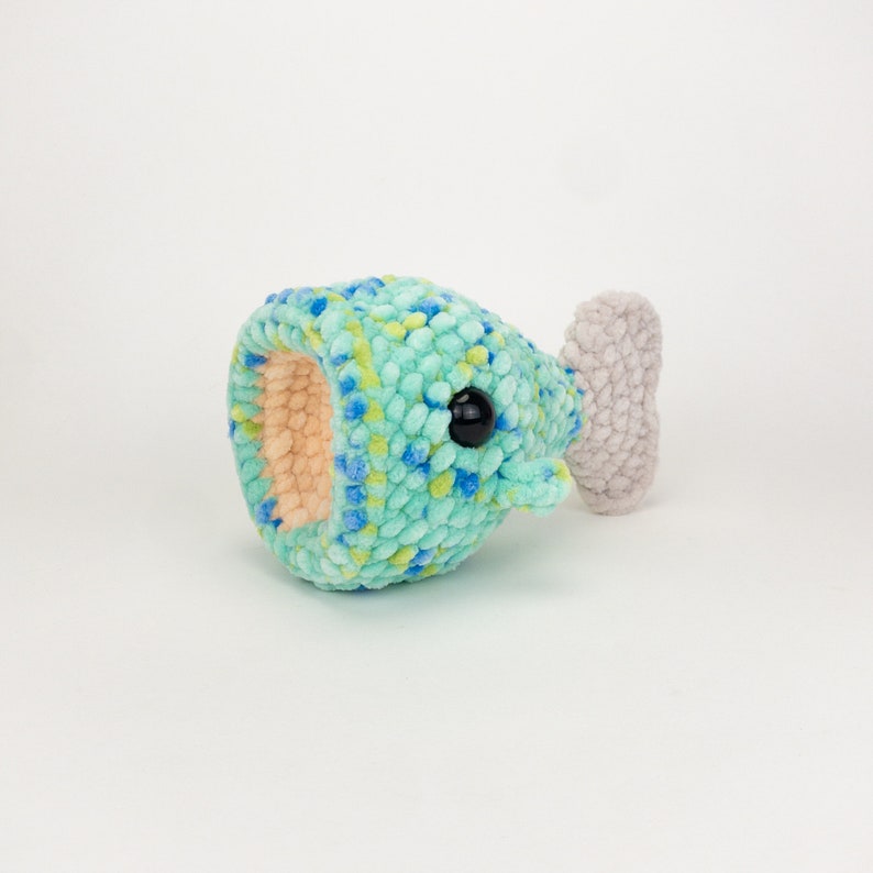 May include: A crocheted blue and green whale plush toy with a gray tail fin and black eyes. The whale is lying on its side with its mouth open.