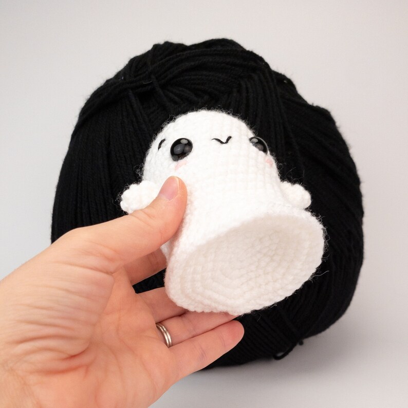 May include: A small, white crocheted ghost with black eyes and a smiling expression. The ghost is held in front of a large ball of black yarn. The ghost has a rounded bottom and small arms.
