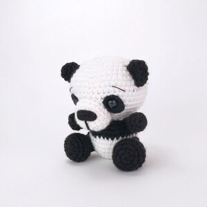 May include: A small crocheted panda bear toy with black ears, eyes, and paws. The panda is sitting with its legs crossed and has a black and white striped body.