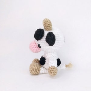Puede incluir: Un juguete de peluche de vaca tejido a crochet con manchas negras, una nariz rosa y orejas marrones. La vaca est&aacute; sentada con la cola enroscada detr&aacute;s de ella.