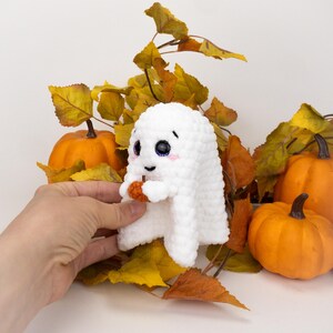 K&ouml;nnte beinhalten: Eine wei&szlig;e geh&auml;kelte Geisterpuppe mit rosigen Wangen und gro&szlig;en Augen, die einen kleinen orangefarbenen K&uuml;rbis h&auml;lt. Der Geist ist von orangefarbenen K&uuml;rbissen und Herbstbl&auml;ttern umgeben und schafft ein festliches Halloween-Thema.