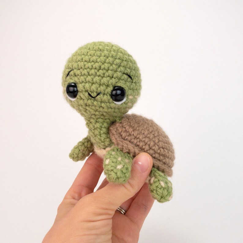 May include: A small green crocheted turtle with brown shell and black eyes. The turtle is being held in a hand.