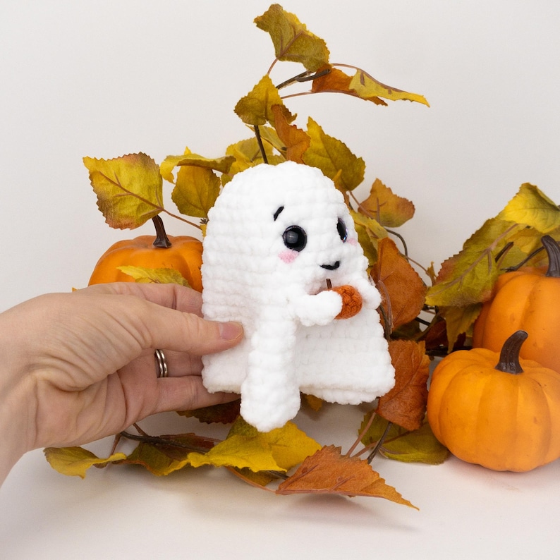 K&ouml;nnte beinhalten: Eine wei&szlig;e geh&auml;kelte Geisterpuppe mit schwarzen Augen und einem kleinen orangefarbenen K&uuml;rbis. Der Geist h&auml;lt den K&uuml;rbis. Der Hintergrund umfasst orangefarbene K&uuml;rbisse und Herbstlaub. Eine niedliche Halloween-Dekoration.
