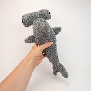 May include: A gray plush hammerhead shark toy with black eyes. The toy is being held by a hand.