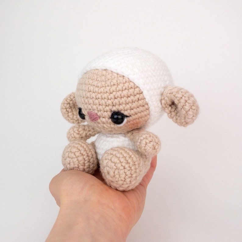 May include: A crocheted lamb toy with a white hat and brown body. The lamb is sitting in a hand.