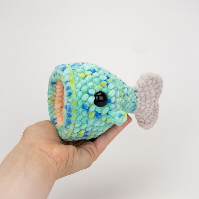 May include: A crocheted plush toy shaped like a fish with a light blue and green body, a gray tail fin, and black eyes.