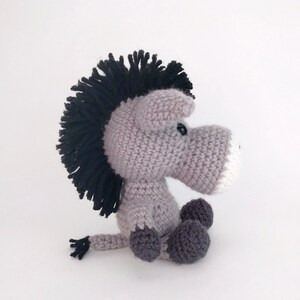 May include: A gray crocheted donkey with a black mane and tail. The donkey has a white nose and black eyes. The donkey is sitting with its legs crossed.
