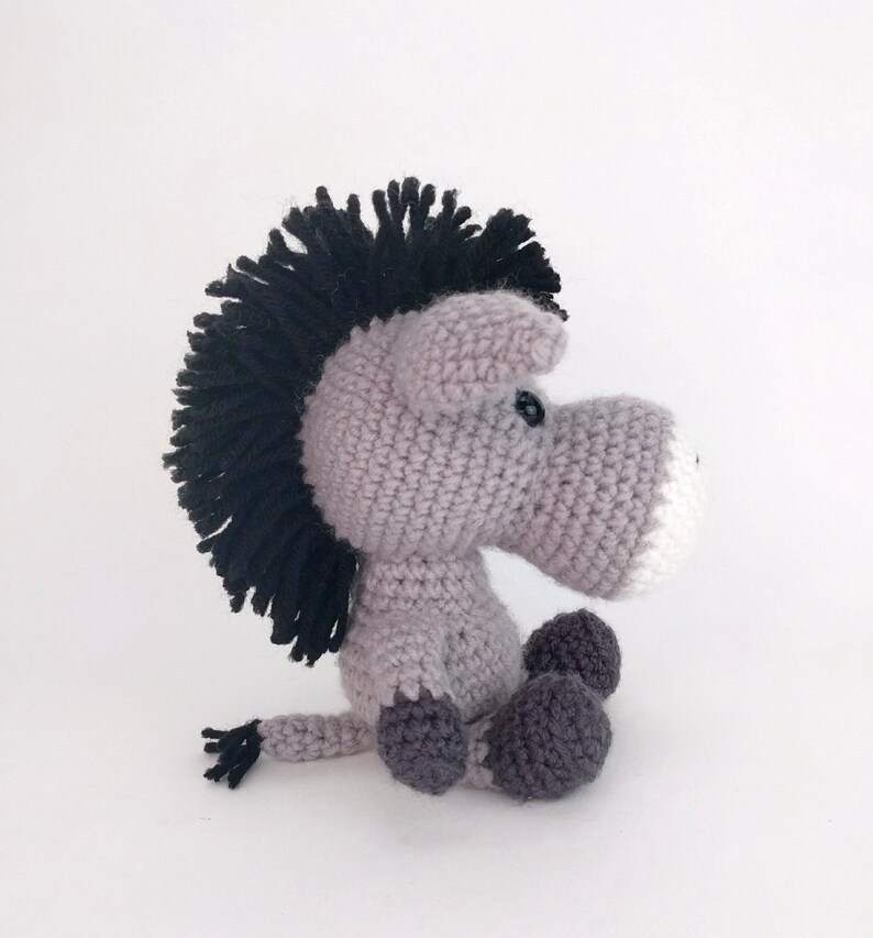 May include: A gray crocheted donkey with a black mane and tail. The donkey has a white nose and black eyes. The donkey is sitting with its legs crossed.
