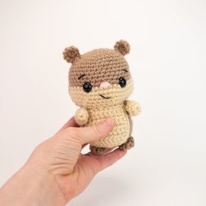 May include: A crocheted brown and beige squirrel plush toy with black eyes and a pink nose. The toy is being held in a hand.