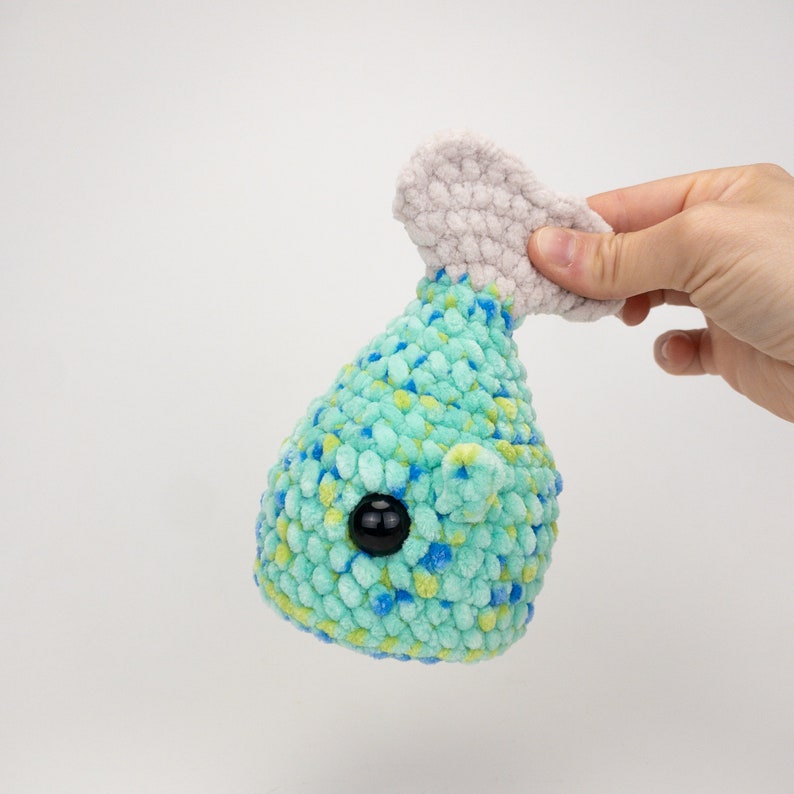 May include: A crocheted blue and green whale plushie with a white crocheted tail fin and a black button eye.