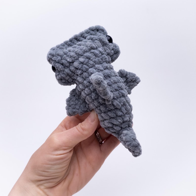 May include: A gray crocheted hammerhead shark plush toy with black eyes. The plush toy is being held in a hand.