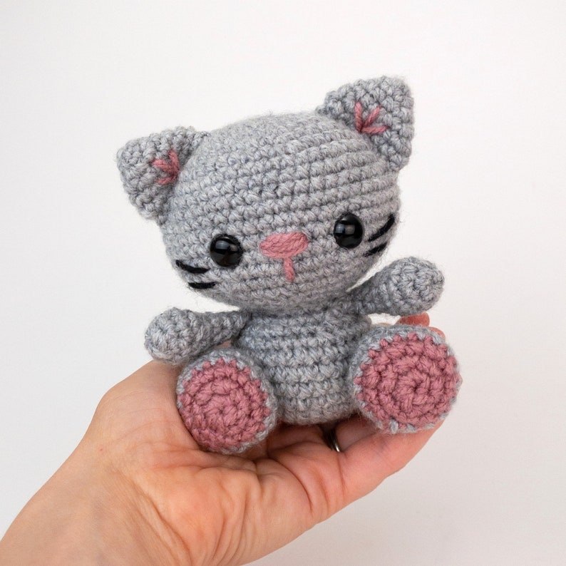 Puede incluir: Un juguete de crochet de gato gris con orejas y patas rosas. El gato tiene ojos bordados negros y una nariz rosa.