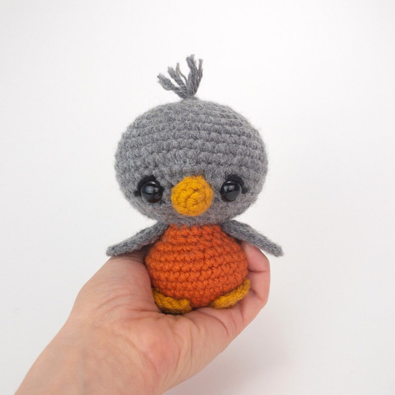 May include: A handmade crochet bird with a gray head, orange body, and small gray wings. The bird has large black eyes and a yellow beak. The bird is a cute, soft toy.