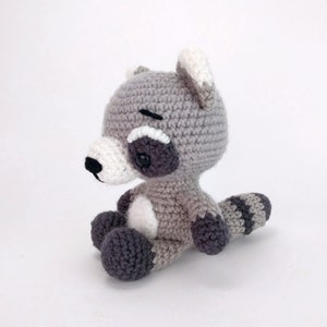 Puede incluir: Un juguete de peluche de mapache tejido a crochet con pelaje gris y blanco, ojos negros y nariz negra. El juguete est&aacute; sentado con la cola enroscada alrededor de sus piernas.
