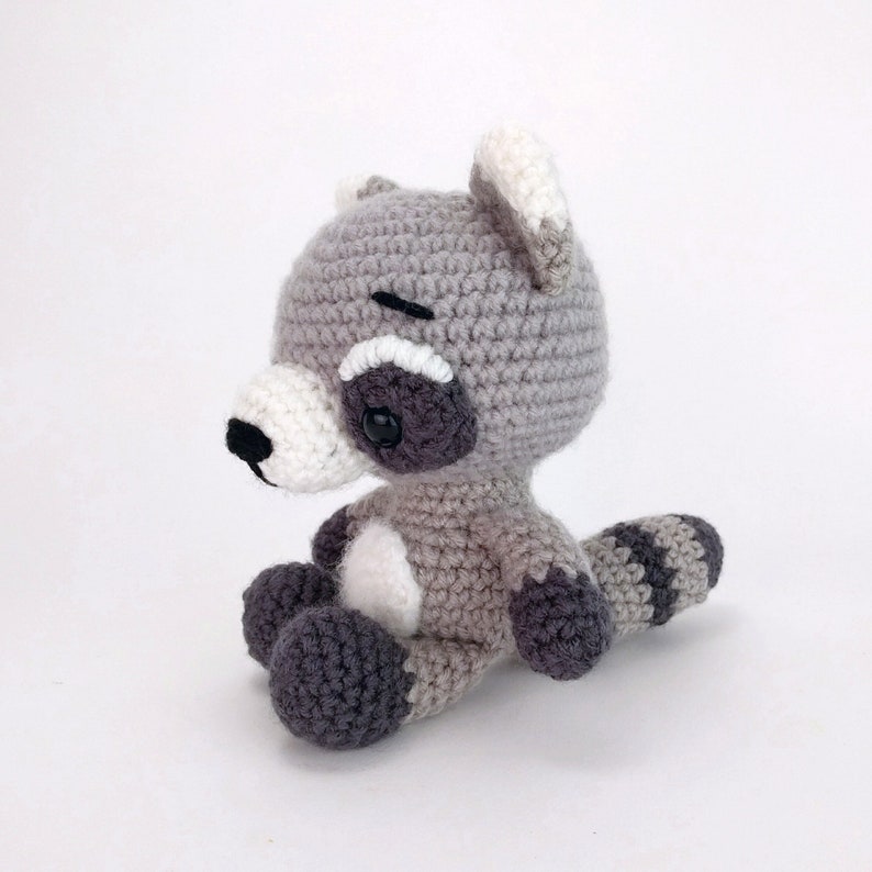 Puede incluir: Un juguete de peluche de mapache tejido a crochet con pelaje gris y blanco, ojos negros y nariz negra. El juguete est&aacute; sentado con la cola enroscada alrededor de sus piernas.