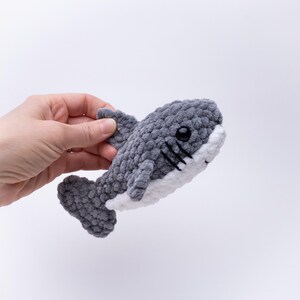 May include: A gray and white crocheted shark plush toy with black eyes. The shark is being held by a hand.