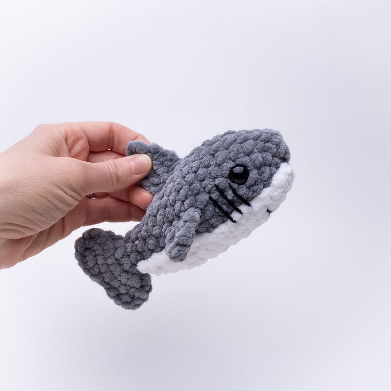 May include: A gray and white crocheted shark plush toy with black eyes. The shark is being held by a hand.