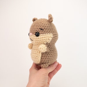 May include: A crocheted brown and beige hamster plush toy with black eyes and a pink nose. The toy is being held in a hand.
