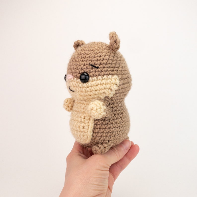 May include: A crocheted brown and beige hamster plush toy with black eyes and a pink nose. The toy is being held in a hand.