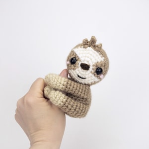 May include: A crocheted sloth toy with brown and cream fur, black eyes, and a white face. The sloth is holding its arms up in the air.