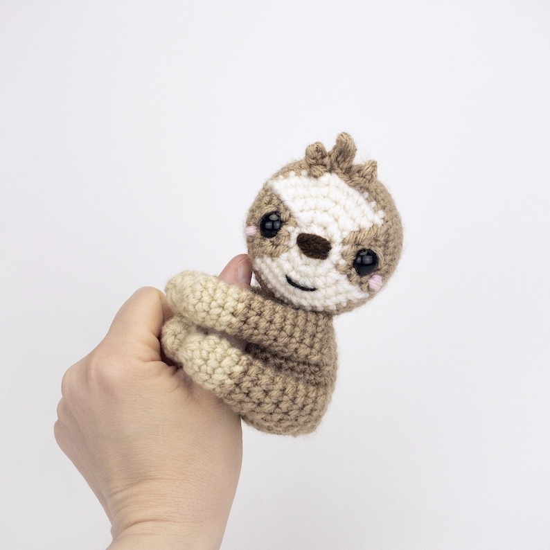 May include: A crocheted sloth toy with brown and cream fur, black eyes, and a white face. The sloth is holding its arms up in the air.
