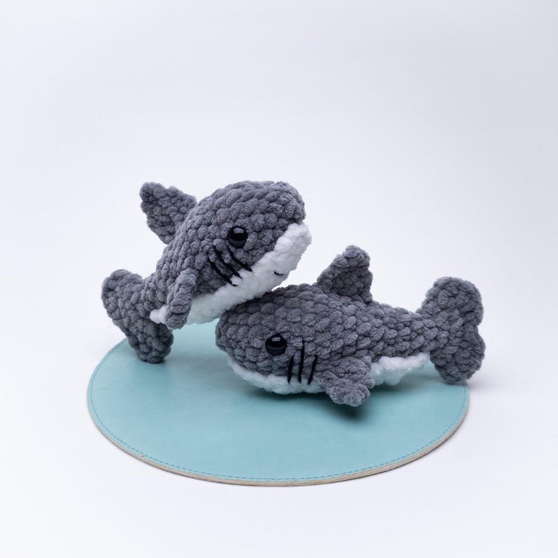May include: Two gray and white crocheted shark plushies with black embroidered eyes and mouths. The plushies are sitting on a light blue round surface.