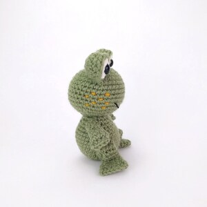 May include: A green crocheted frog toy with black eyes and yellow spots. The frog is sitting on a white surface.