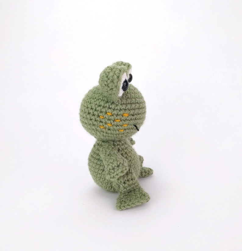 May include: A green crocheted frog toy with black eyes and yellow spots. The frog is sitting on a white surface.