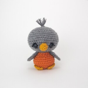 May include: A handmade crochet bird with a gray head, orange body, and yellow feet. The bird has large black eyes and a small yellow beak. A small tuft of gray yarn sits atop the head. The bird is a cute, whimsical design.