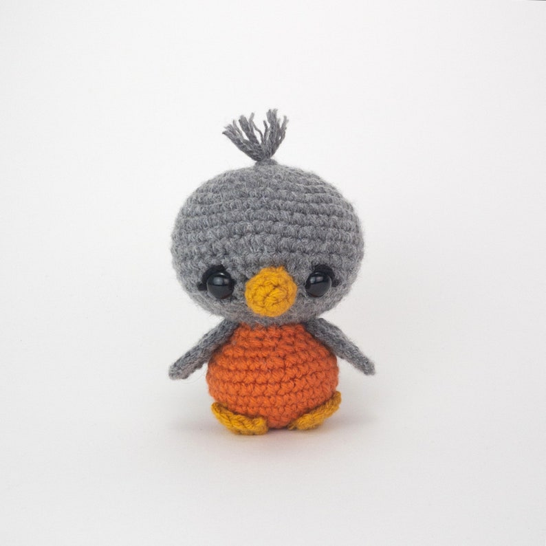May include: A handmade crochet bird with a gray head, orange body, and yellow feet. The bird has large black eyes and a small yellow beak. A small tuft of gray yarn sits atop the head. The bird is a cute, whimsical design.